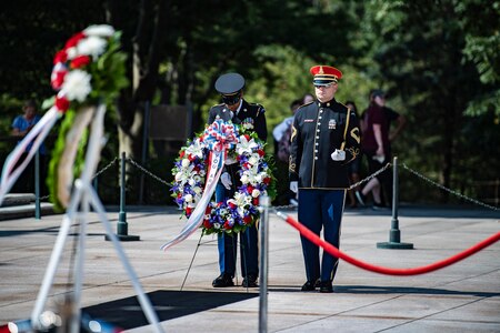A soldier is adjusting a wreath for a presentation at the Tomb of the Unknown Soldier as another soldier holding a bugle stands next to him. There is another colorful wreath that as already been placed near the Tomb of the Unknown Soldier in the foreground.