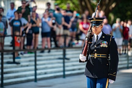 Woman in Army dress uniform marches with a rifle and bayonet on her shoulder as people in the background watch.