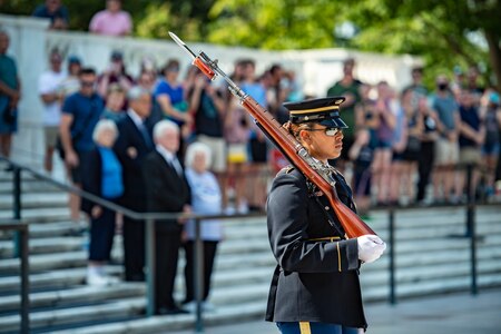 Woman in Army dress uniform and sunglasses holding a large rifle and bayonet over her shoulder while facing away from the crowd of people on the white marble steps behind her. She is guarding the Tomb of the Unknown Soldier at Arlington National Cemetery