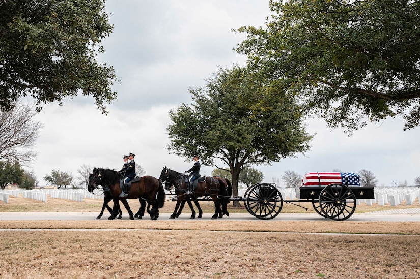 Soldiers on horses draw a flag-draped casket along a road in a cemetery with trees and headstones in the backdrop.