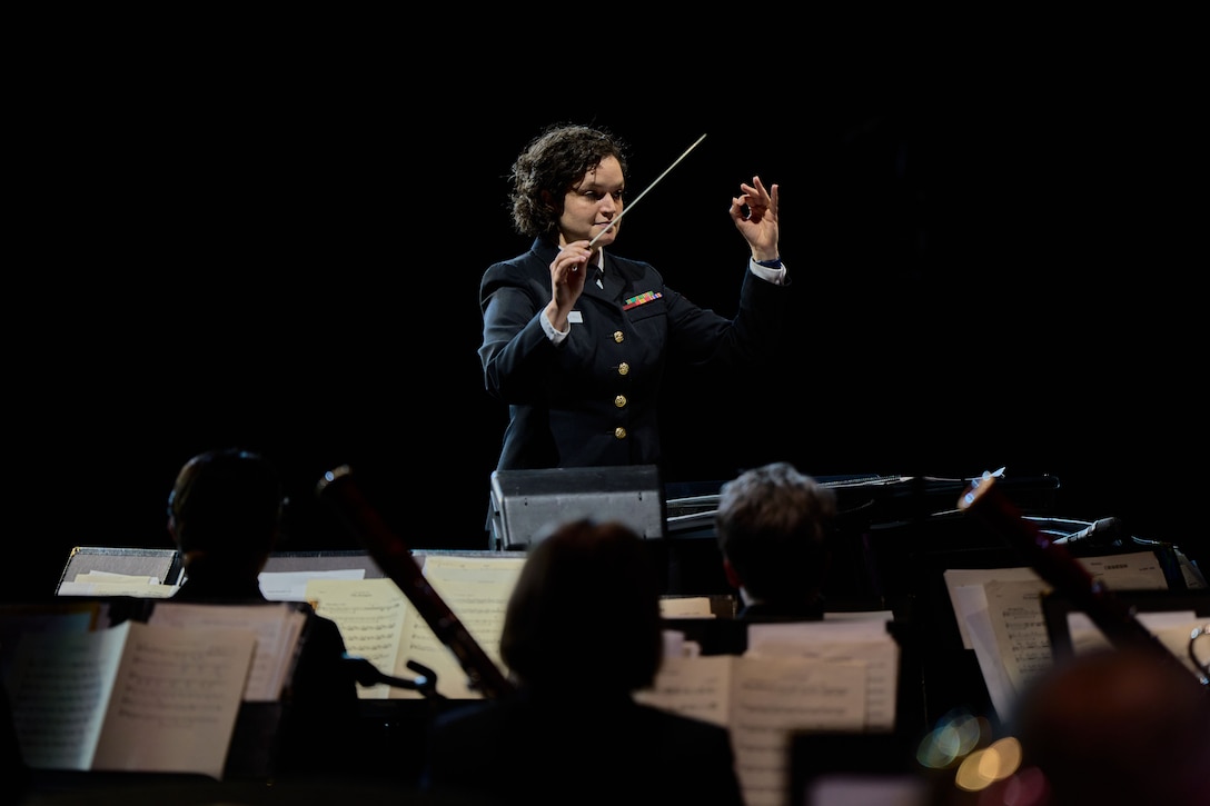 A sailor in dress uniform on a darkened stage, motions with a hand and a baton before an orchestra.
