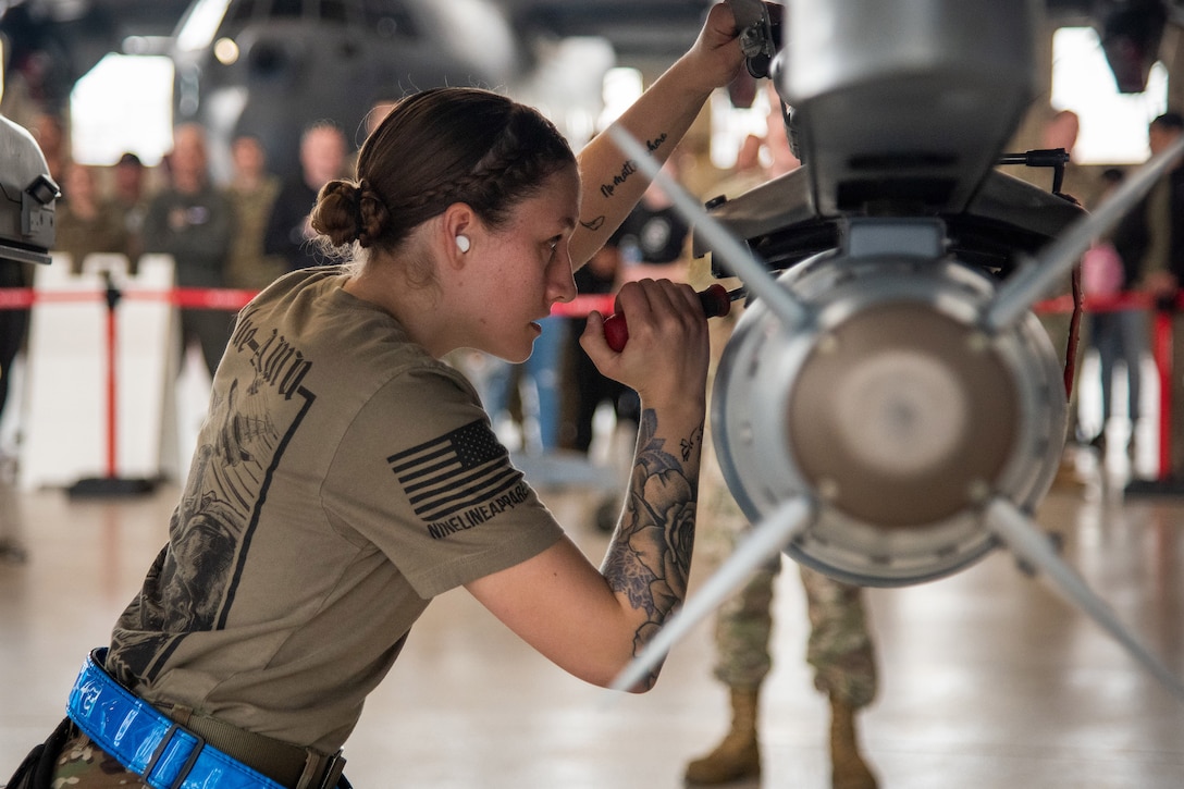 An airman secures an ordnance suspended from an aircraft while a group looks on in the background.