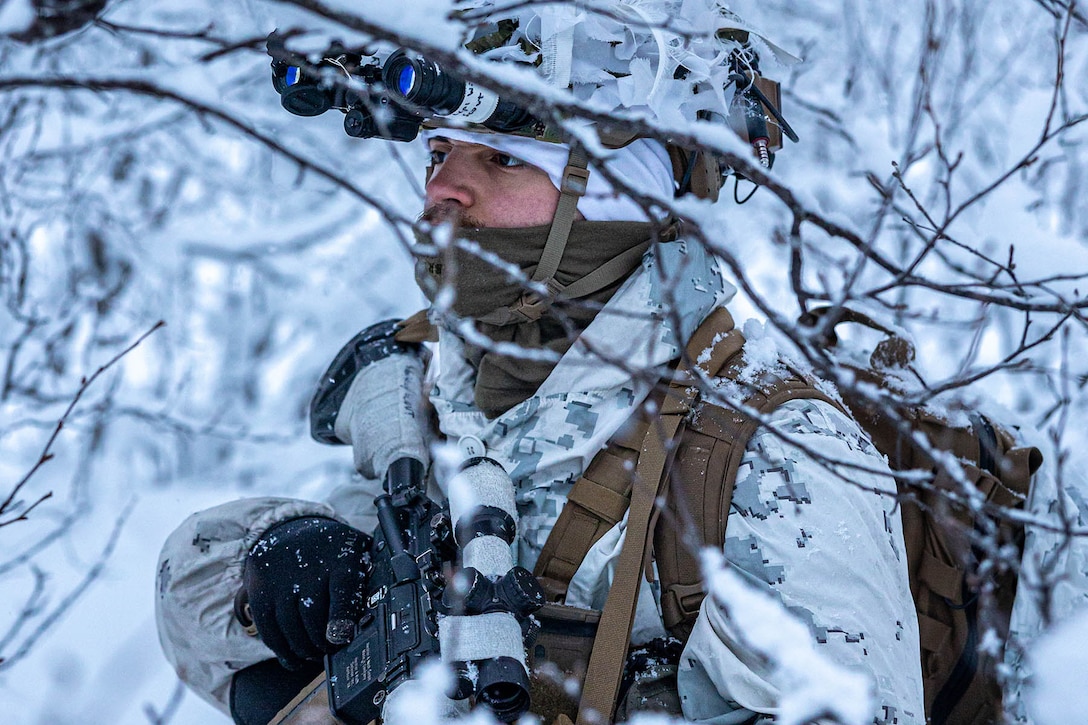 A Marine wearing white camouflage winter gear holds a weapon while standing in a snow-covered, forested area.