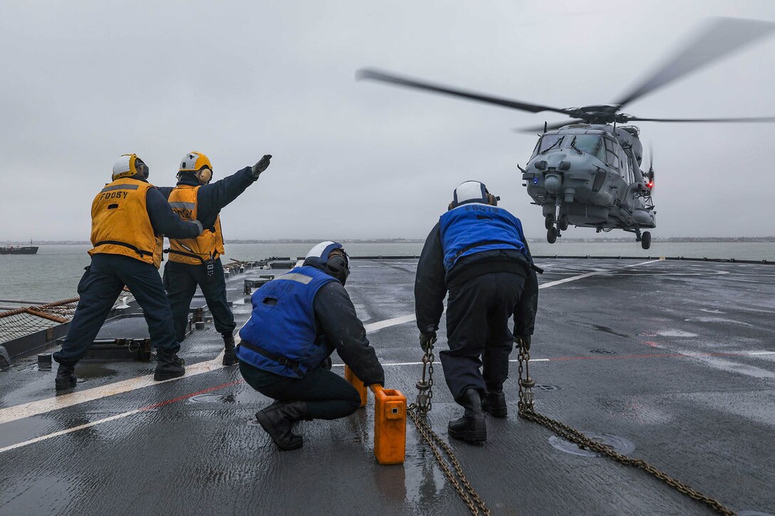 Two sailors in yellow shirts and two in blue position themselves on a ship's flight deck as a helicopter approaches to land.