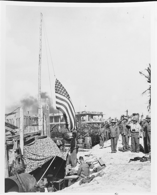 Fourth division Marines salute as the flag is raised on Roi Island, Kwajalein Atoll, marking the capture of this Japanese stronghold, 2 February 1944. Work goes on in the command post at the base of the flagpole. In the background is the remains of a three story concrete building, victim of the pre-invasion bombardment. Naval History and Heritage Command Photo.