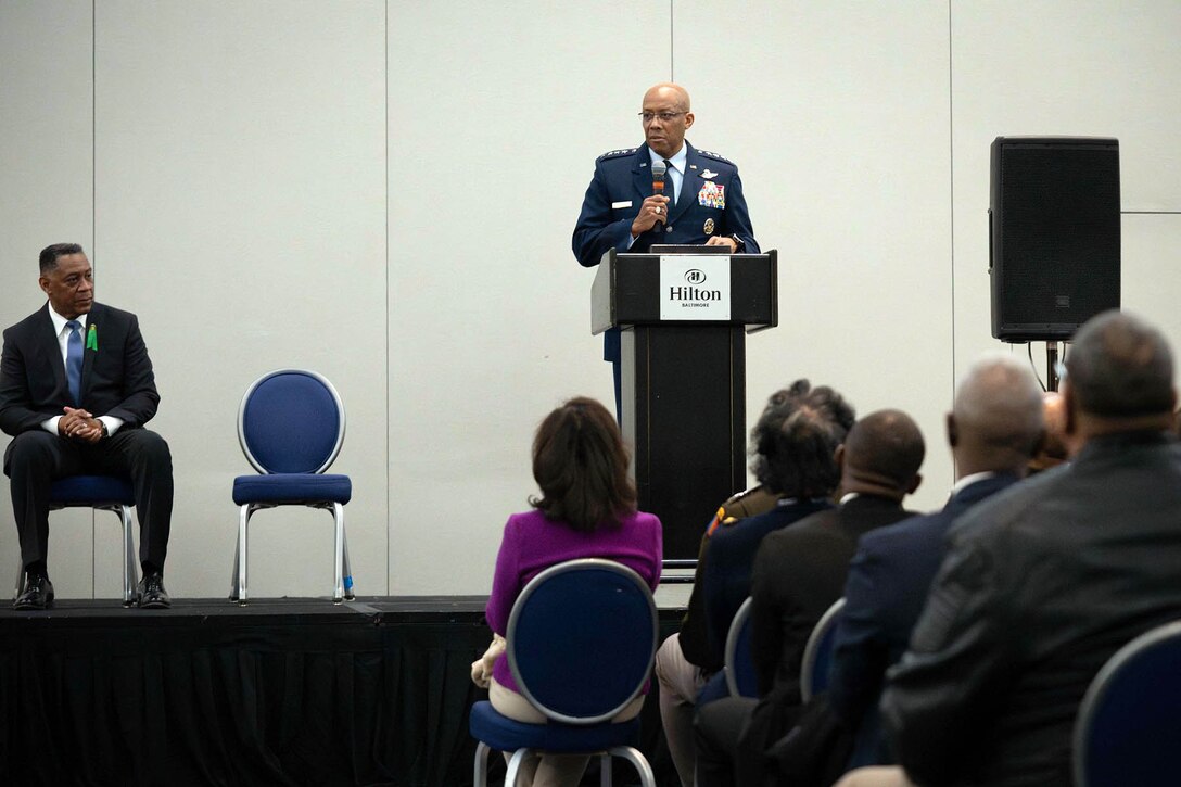 Air Force Gen. CQ Brown, Jr. speaks at a lectern on a stage where a civilian is sitting, facing a seated audience.