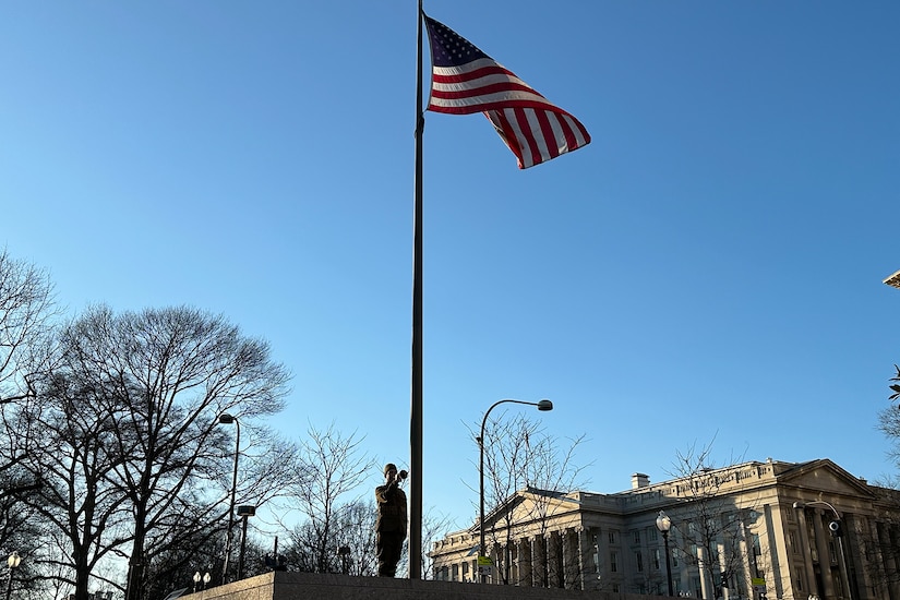 A person wearing World War I-era uniform stands at attention under a U.S. flag with a bugle raised to their mouth.