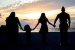 Four people in silhouette hold hands as they look at the setting sun.