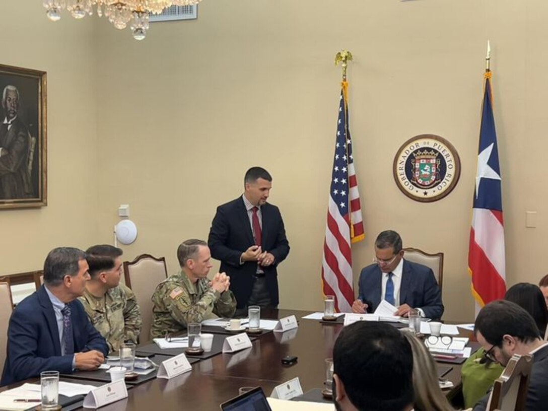 Puerto Rico Governor Pedro Pierluis seated at a table with Col. Charles decker and another soldier dressed in camouflage and others dressed in suits.