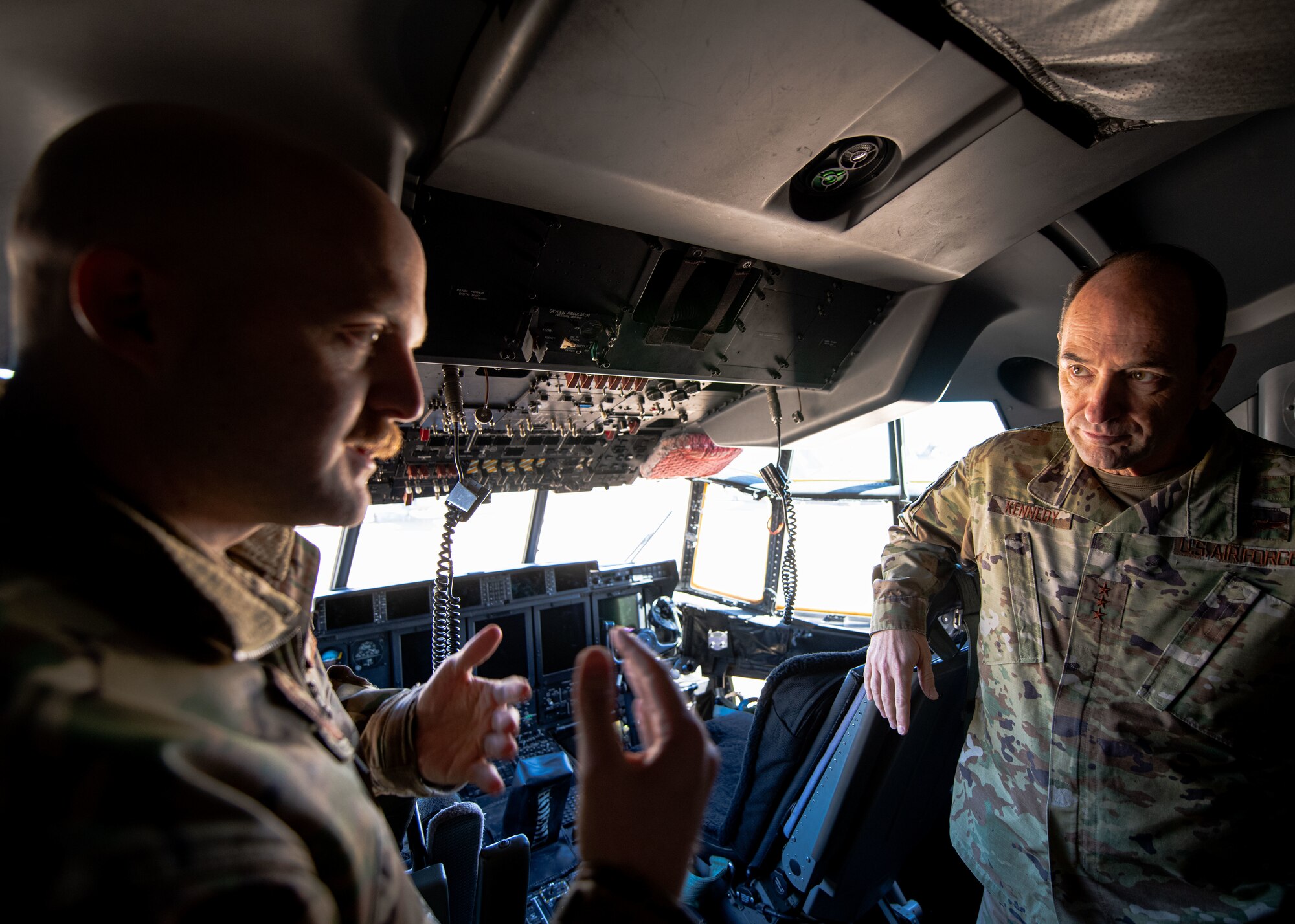 A man in military uniform speaks to another man in military uniform while inside a military aircraft cockpit.