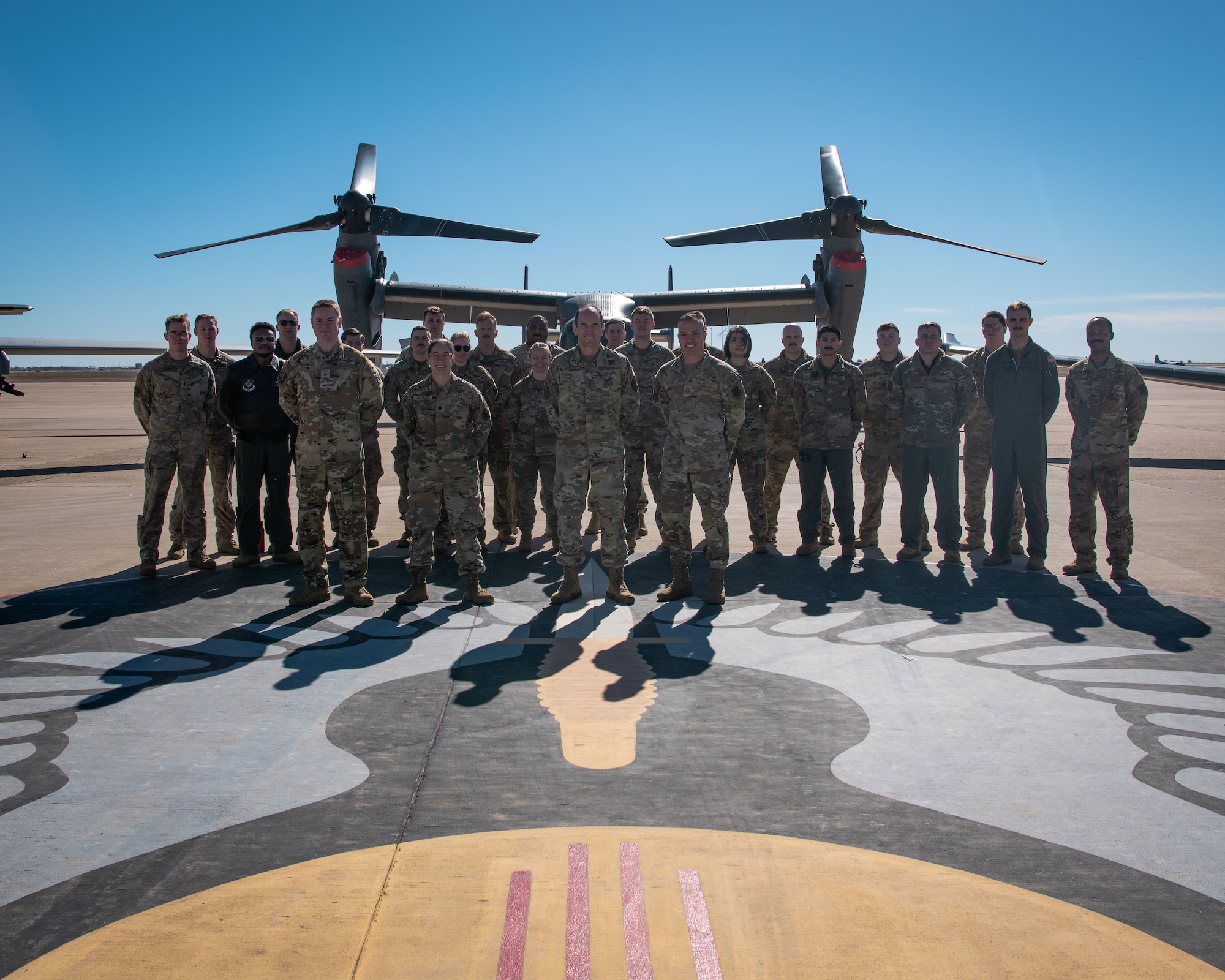 A group of people in military uniform pose for a photo in front of a military tiltrotor aircraft on a tarmac.