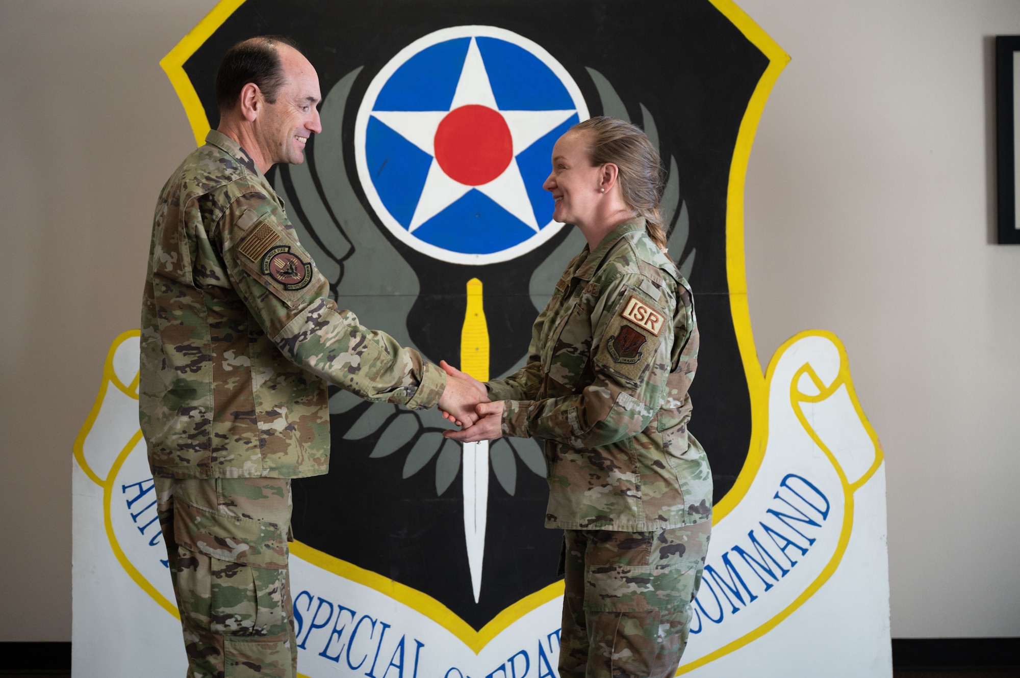 A man and woman in military uniform shake hands in front of a large military logo for Air Force Special Operations Command.