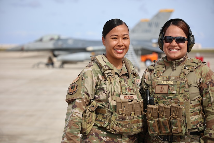 Two airmen stand and smile for a photo on a flight line, with a jet parked in the background.