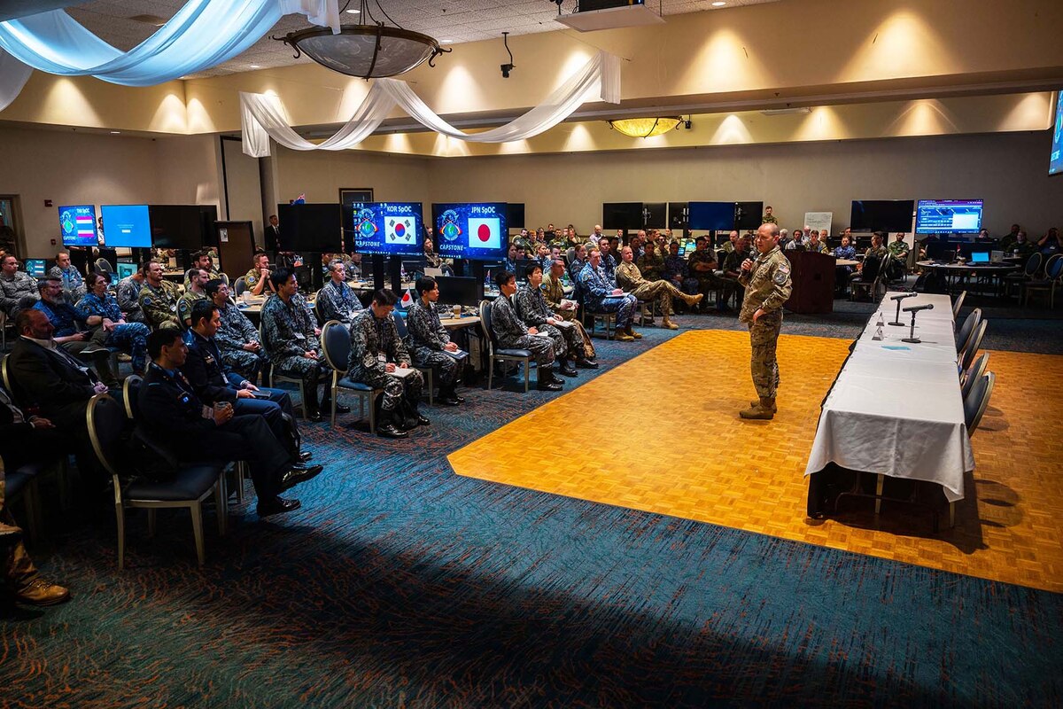 A person in uniform stands with a microphone in his hand as he addresses seated members of foreign militaries in uniform.