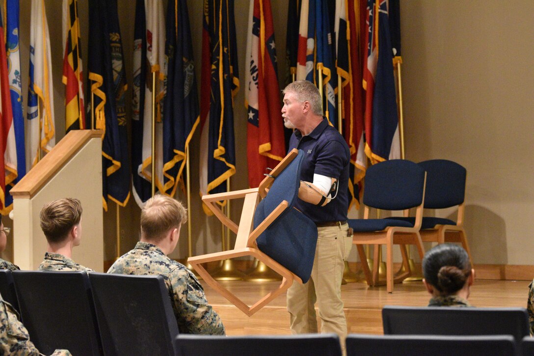 Lee Shelby holds up a chair, demonstrating the capabilities his prosthetics give him, in front of a group of an audience at Combined Arms Training Center, Camp Fuji, Japan, February 16, 2024. (U.S. Marine Corps photo by Song Jordan)