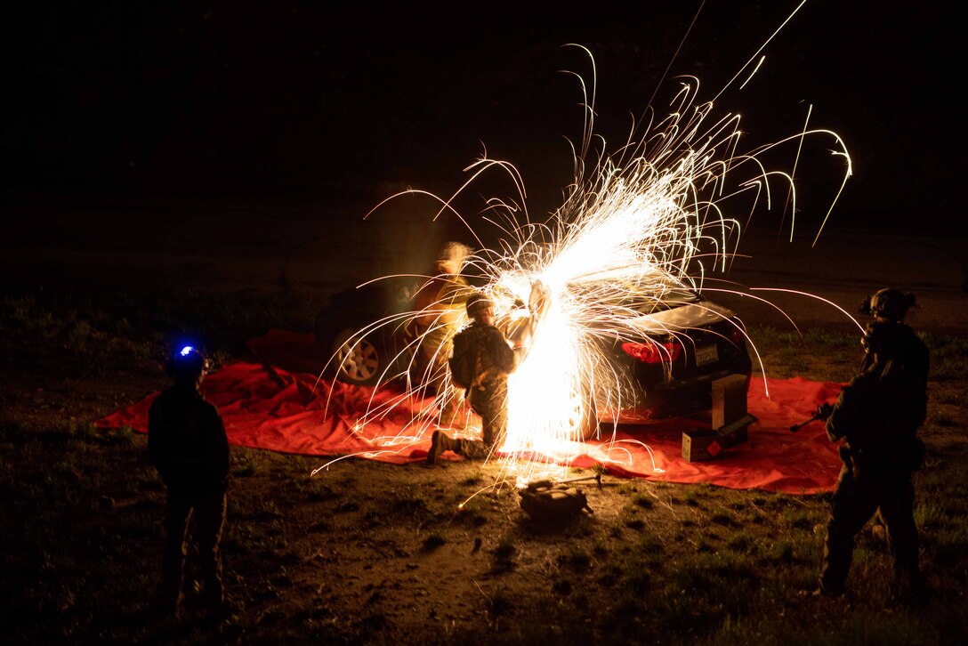 Sparks fly as a Marine uses a saw to cut through metal during a night exercise. Other Marines stand and watch.