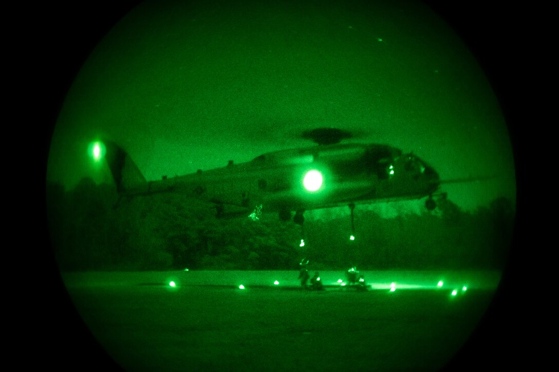 Marines, illuminated by a green light, attach an I-beam to a military helicopter during nighttime training.