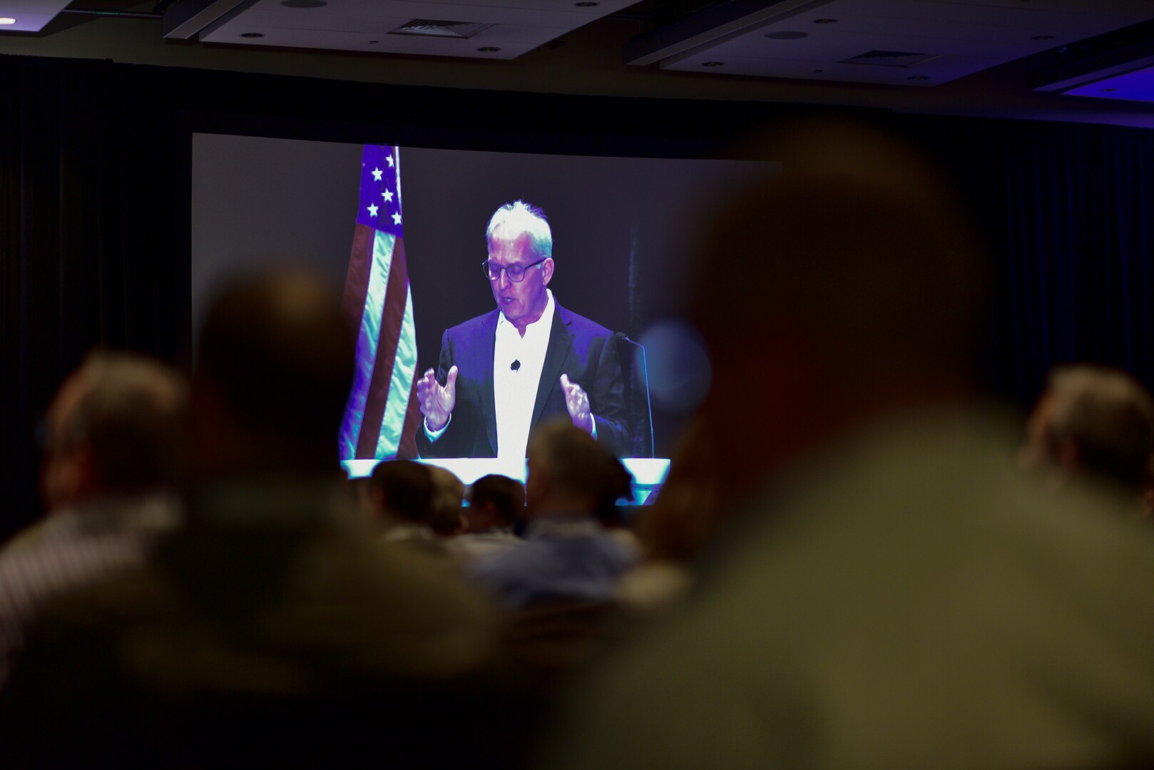 A video of a person in a business suit is projected on a screen before an audience.