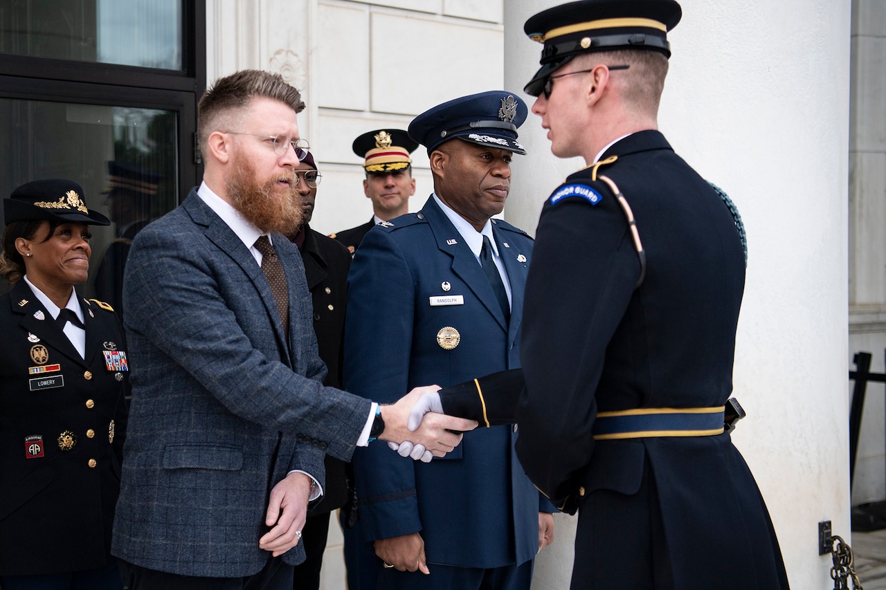 Uniformed service members and a civilian in a business suit greet one another.