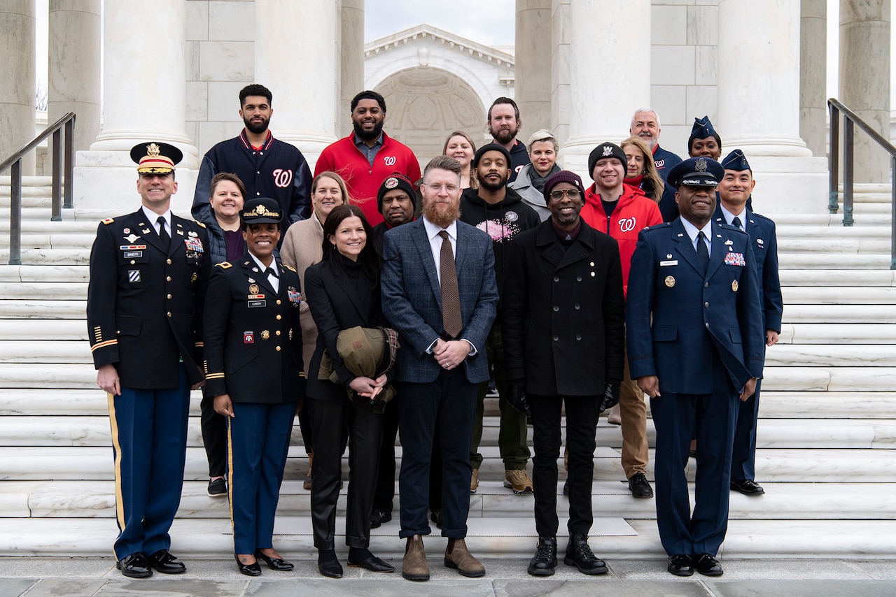 A group of professional baseball players and armed forces senior leaders stand on steps outdoors.