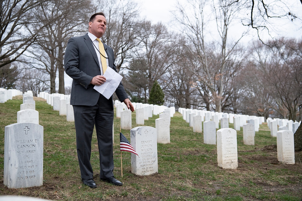 A person in a business suit motions toward a military grave marker.