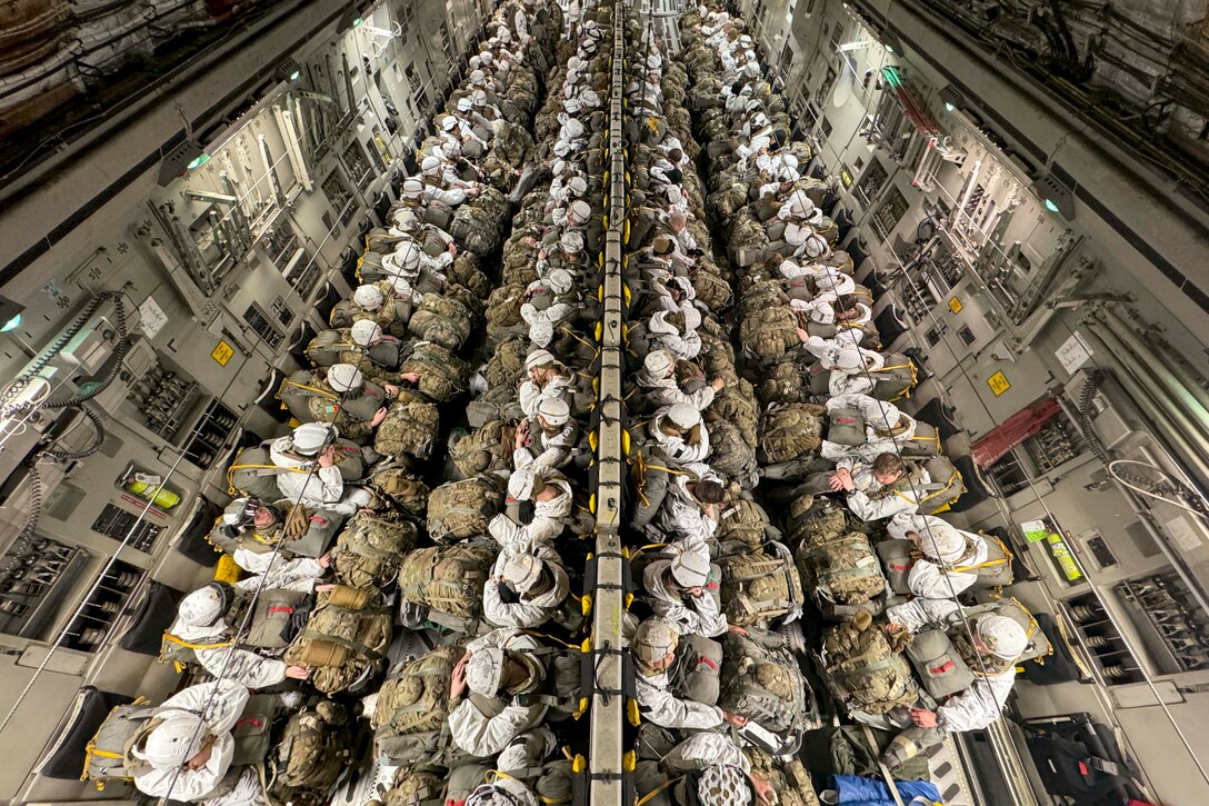 Dozens of soldiers sitting in an aircraft wearing parachute gear carry backpacks in their laps as seen from above.