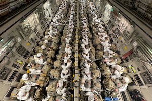 Dozens of soldiers sitting in an aircraft wearing parachute gear carry backpacks in their laps as seen from above.