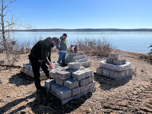 Two men in hats and green jackets stand near four piles of concrete blocks with dirt in the foreground and a lake in the background.