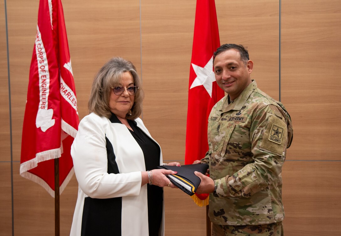 Army soldier in camouflage uniform presenting a folded black flag to a lady wearing a black and white blouse in front of 2 red flags.