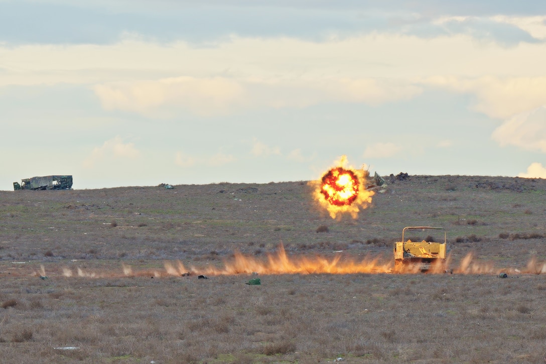 A projectile explodes in midair above a large field with a line of flame and a vehicle in the foreground and another vehicle on the horizon.