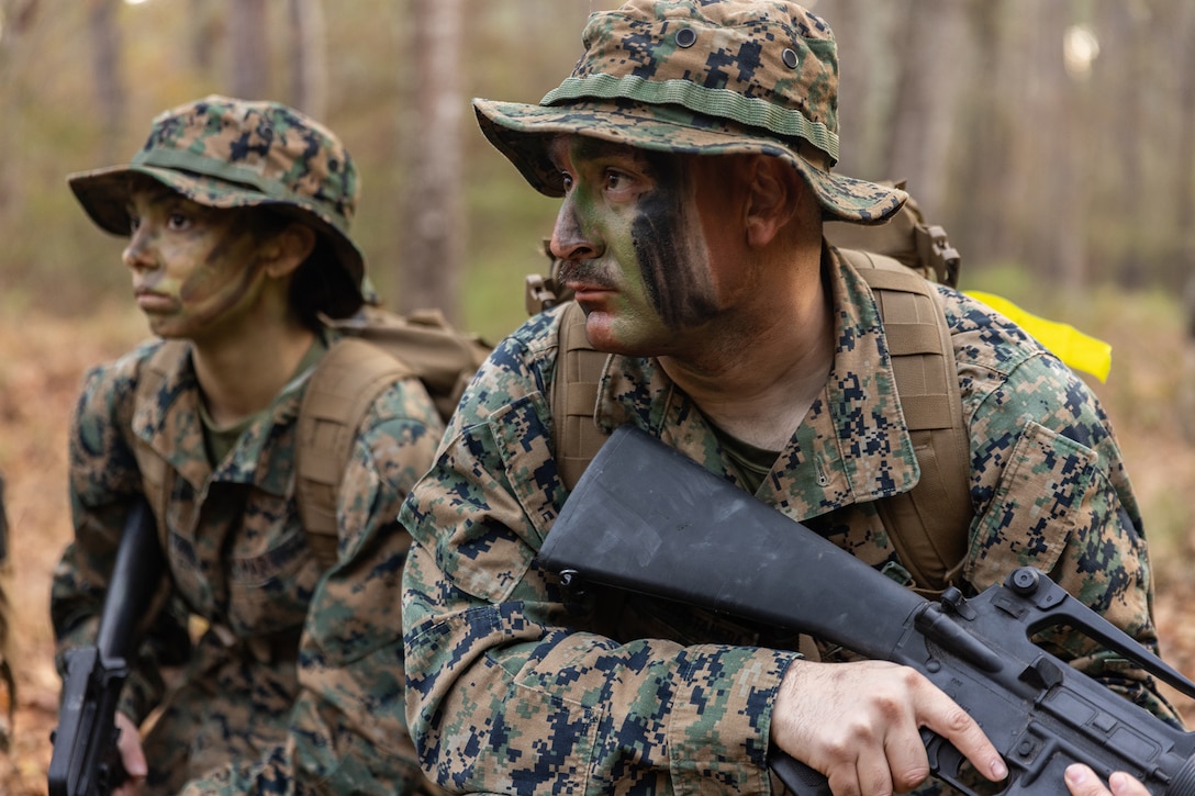 Close-up of two uniformed Marines wearing camouflage face paint and holding weapons in a wooded area.