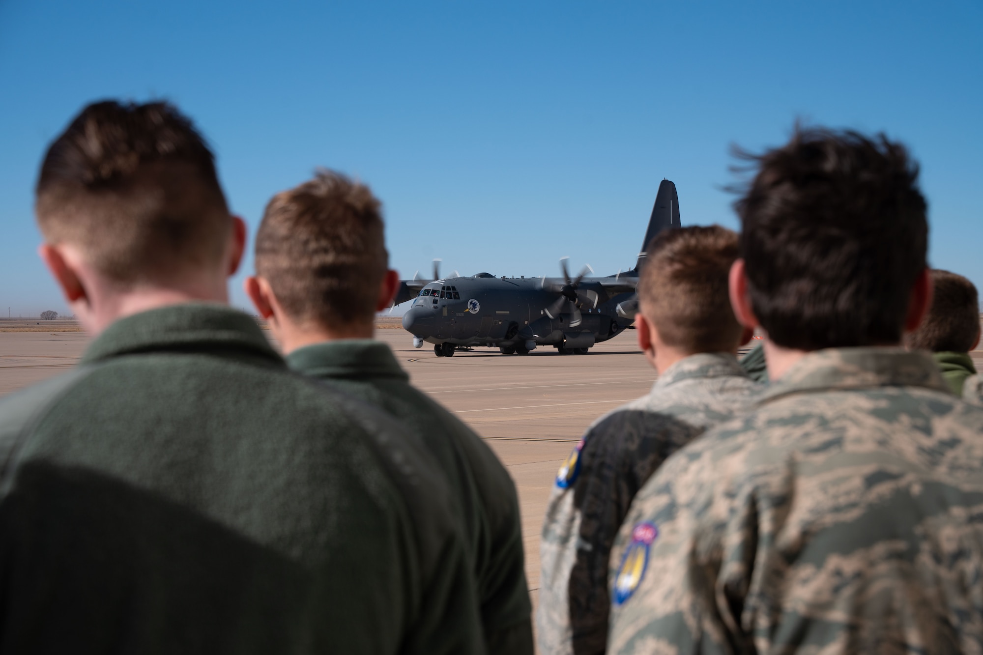 A group of young adults in military uniform stand outside while watching a military aircraft taxi.