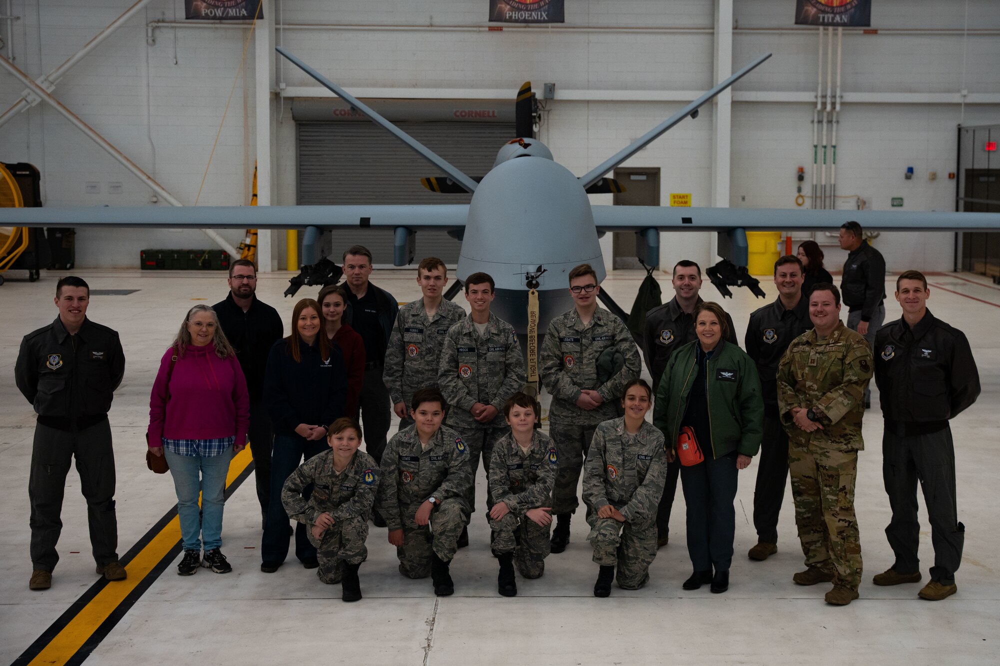 A group of adults and young adults in a mixture of military uniform and casual civilian attire pose in front of a military aircraft while inside an aircraft hangar for a group photo.
