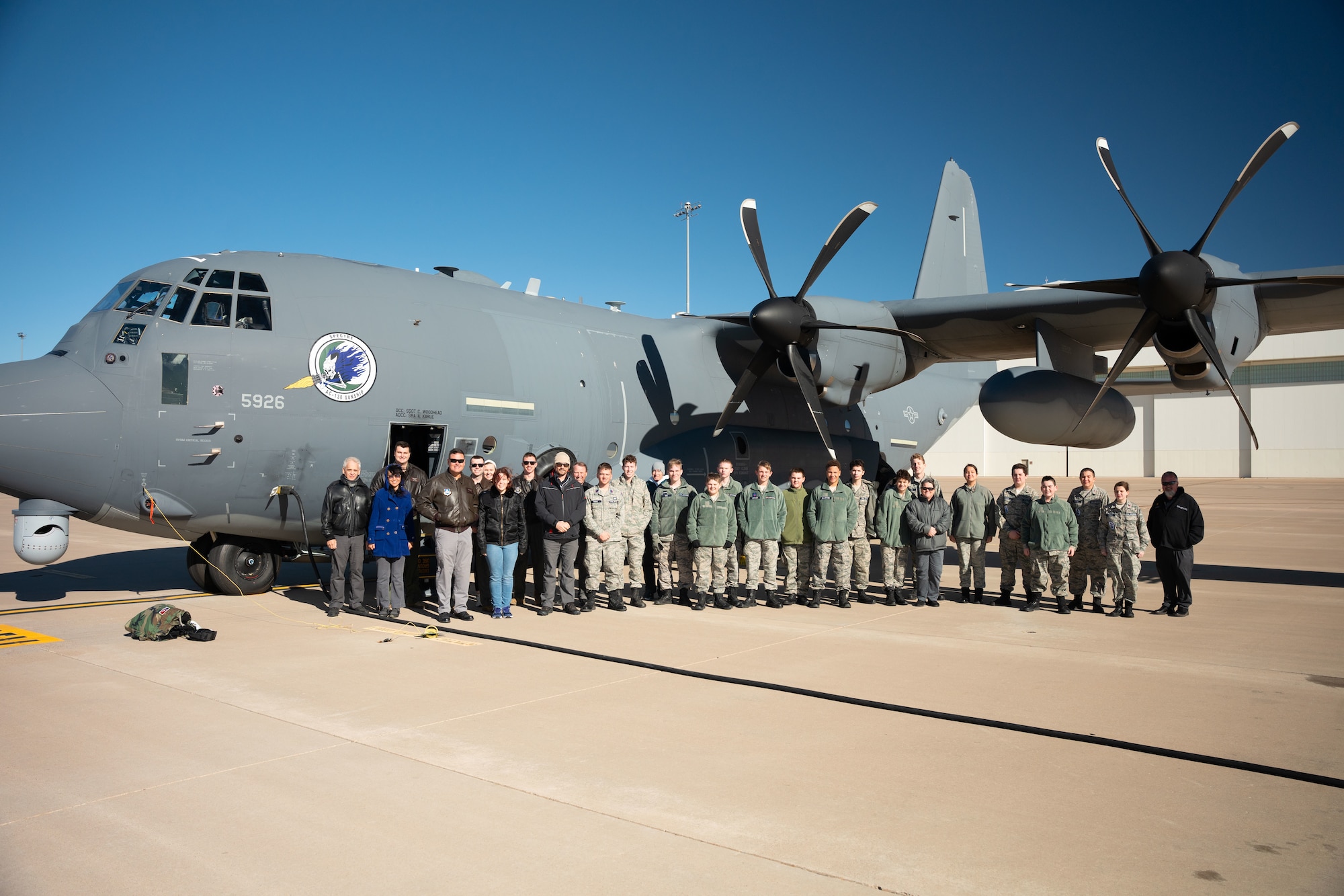 A group of young adults and adults in a mixture of military uniform and casual civilian attire stand in front of a military aircraft outside for a group photo.