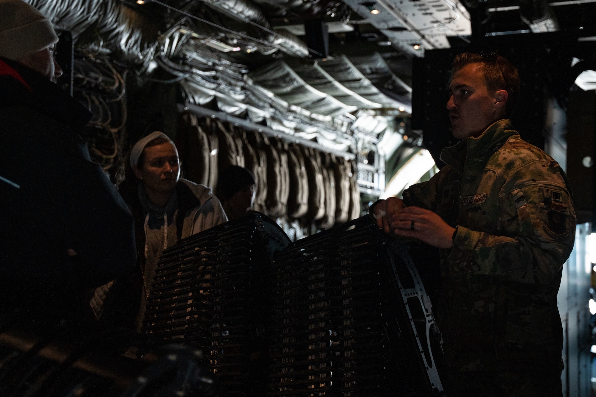 A man in military uniform speaks to a group of young adults in military uniform while holding a piece of equipment inside a military aircraft.