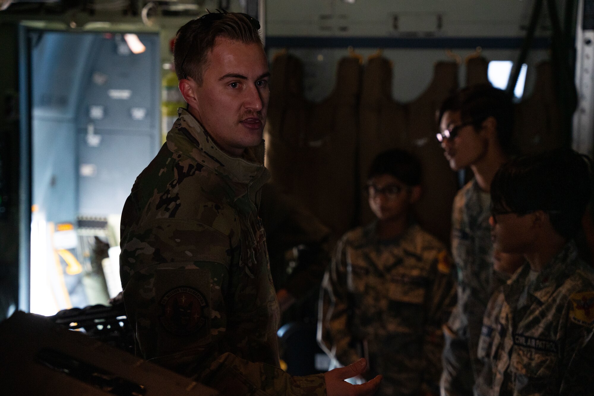 A man in military uniform briefs a group of young adults in military uniform while inside a military aircraft.
