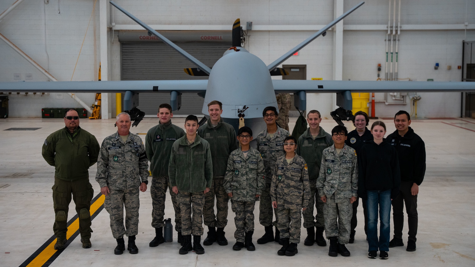 A group of adults and young adults in a mix of military uniform and casual civilian attire stand in front of a military aircraft for a group photo inside an aircraft hangar.