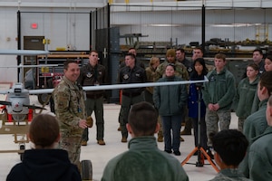 A man in military uniform speaks to a group of men and women in military uniform in front of a small aircraft inside a hangar.