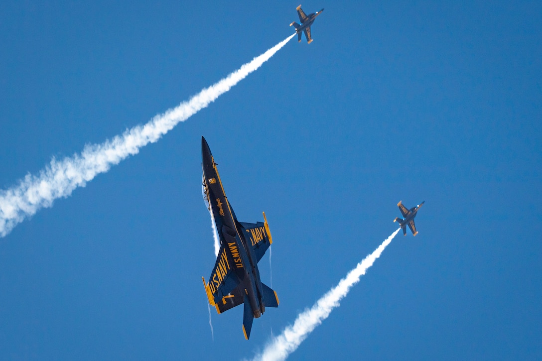 Two aircraft leave streaks in the sky while flying in formation to the right as another aircraft flies between their streaks to the left.