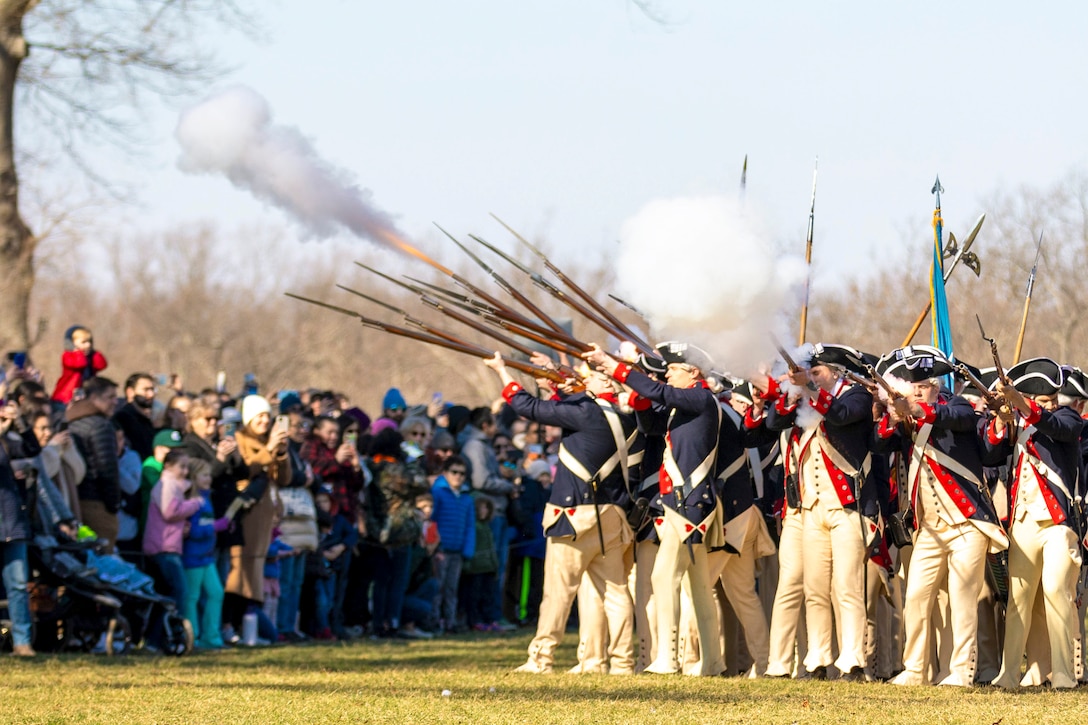 Soldiers in ceremonial uniforms gather in a circle and fire weapons in a field as people watch.