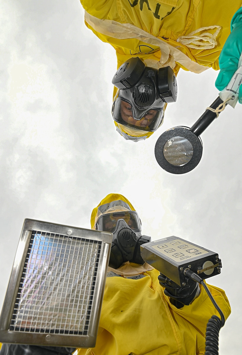 Two Airmen wearing yellow protective gear use tools to simulate decontamination procedures.