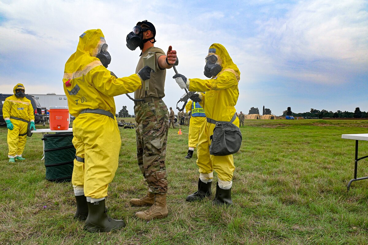 Two Airmen are wearing protective equipment are simulating decontaminating an Airman wearing a gas mask using sprayer tools in a field.