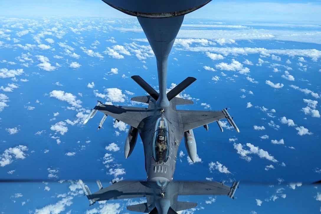 Part of an F-16 is seen in a reflection as it approaches a larger aircraft in a blue sky with small clouds.