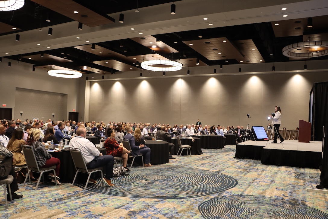 A conference room filled with people watching a speaker talk on a platform stage.