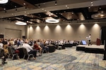 A conference room filled with people watching a speaker talk on a platform stage.