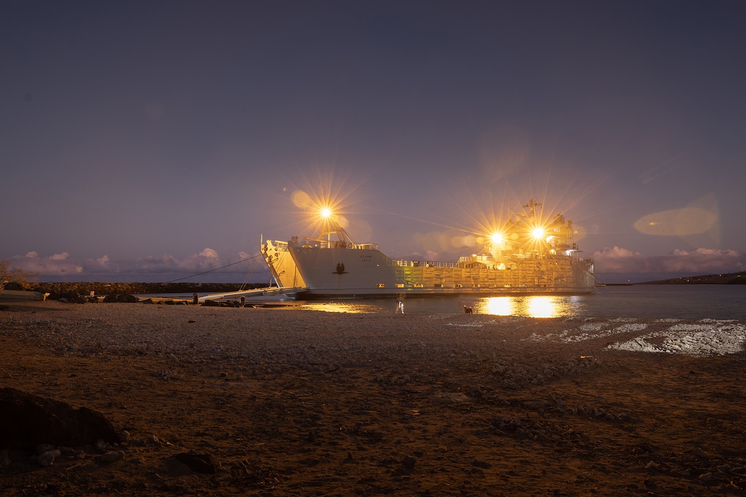 A military vessel, docked at a shoreline, prepares to receive cargo at twilight.