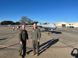 Two men in military flight suits stand posed in front of an remotely piloted aircraft.