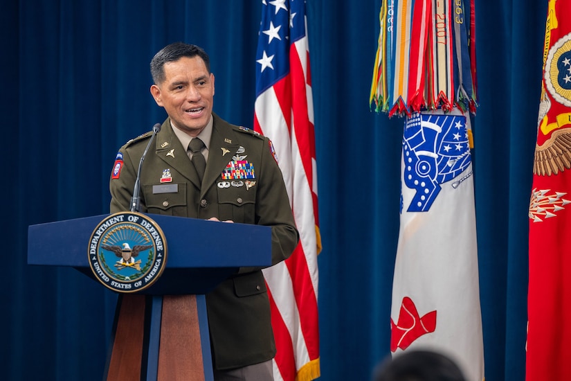 A solider stands at a lectern adorned with a DOD seal in front of flags on a stage.