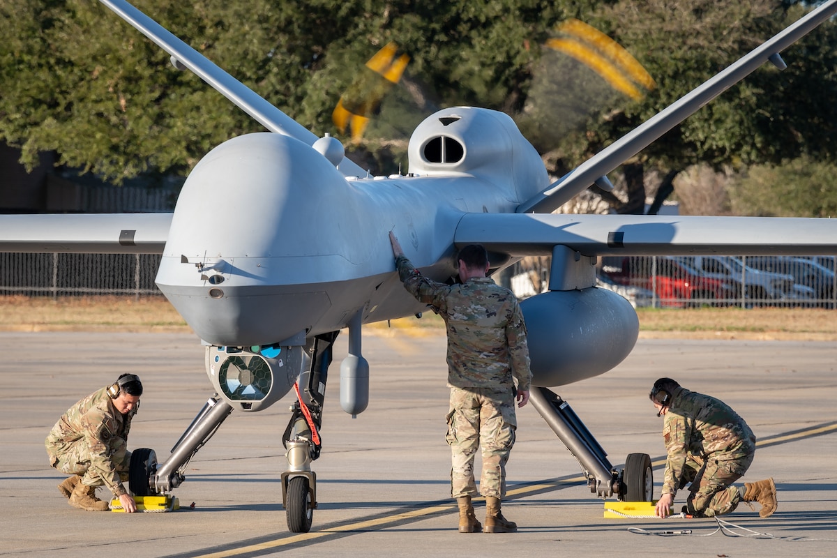 Two men in military uniforms put chocks on the wheels of a remotely piloted aircraft while a third places a hand on the aircraft.