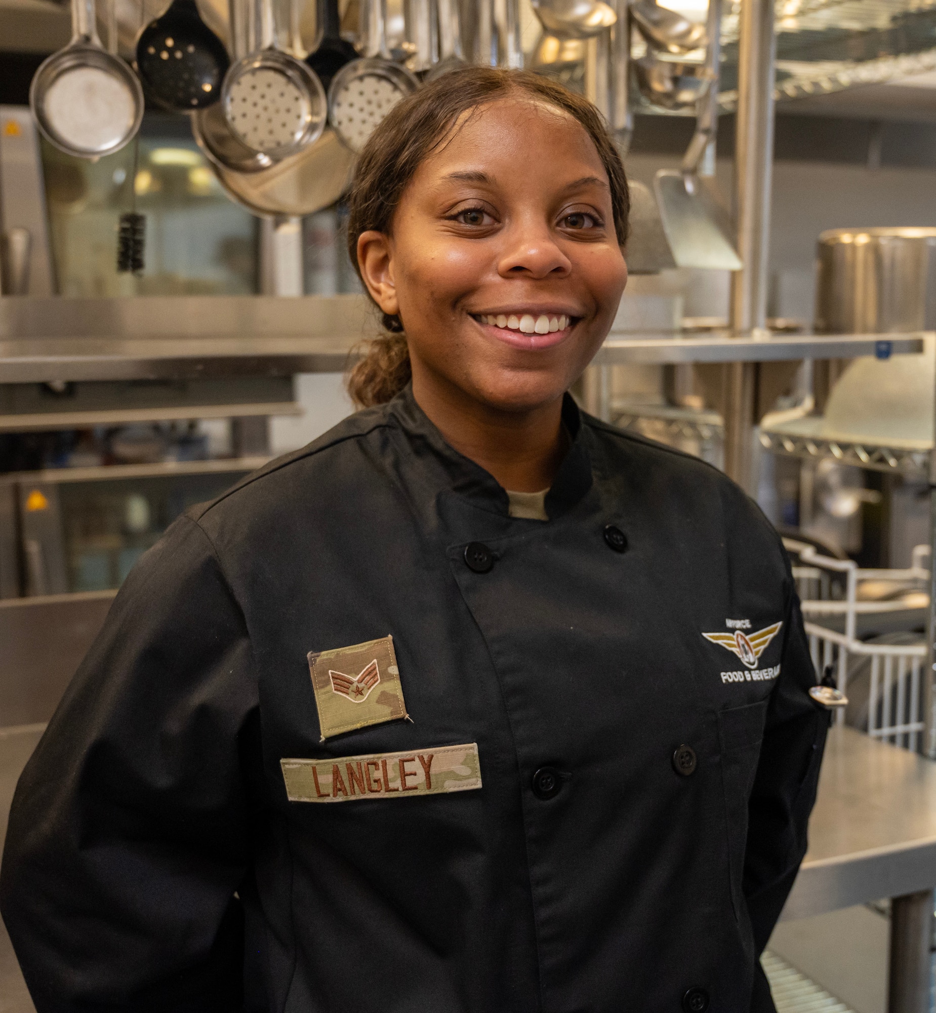A U.S. Air Force Airman proudly poses for a photo in a military kitchen. She is wearing the new Services "dress black" chef's uniform. In the background, ladles, pots and pans hang from an overhead rack.